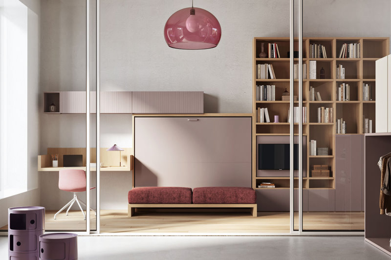 Piccolo living in legno e finitura rosa con letto trasformabile chiuso, scrivania sospesa, grande libreria asimmetrica a parete